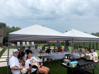 Backyard pool party with family and friends seated under white canopy tents at checkered tables, outdoor griddle and food on the table, cloudy sky over a suburban lawn.