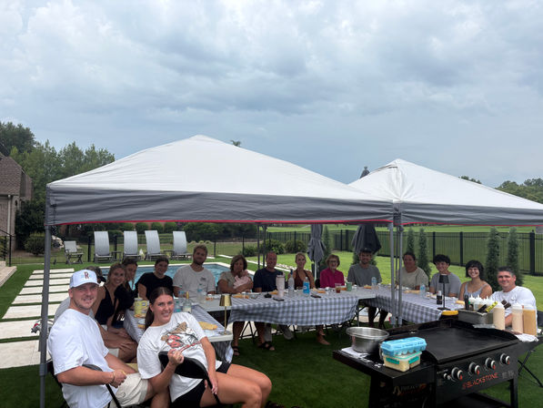 Backyard pool party with family and friends seated under white canopy tents at checkered tables, outdoor griddle and food on the table, cloudy sky over a suburban lawn.
