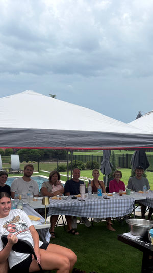 Backyard pool party with family and friends seated under white canopy tents at checkered tables, outdoor griddle and food on the table, cloudy sky over a suburban lawn.