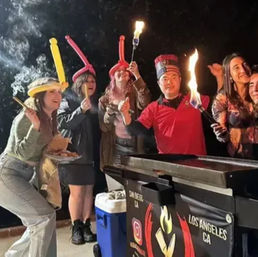 Festive nighttime street-food scene with a smiling group around a food cart holding flaming skewers and wearing colorful balloon hats — urban outdoor celebration.