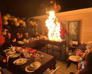 Outdoor nighttime backyard deck party with guests seated at long tables, black-and-gold balloon garland, and a hibachi grill shooting a dramatic tall flame.