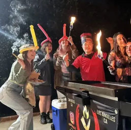 Nighttime Los Angeles street food scene: group wearing balloon hats posing by a cart, holding flaming skewers and smiling.