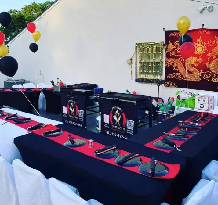 Outdoor hibachi-style catering setup for an Asian-themed party with U-shaped black-and-red tables, place settings and chopsticks, tabletop grills in the center, gold and black balloons and a red dragon wall hanging.