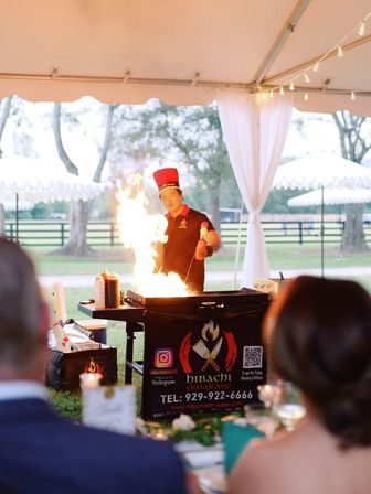 Hibachi chef in a red hat creating a dramatic flame on a teppanyaki grill under a white event tent at an outdoor wedding reception on a grassy lawn, guests watching the live cooking show.
