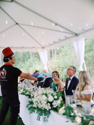 Playful chef in a red hat aims a blue blaster at laughing guests seated at the head table of an outdoor wedding reception tent, surrounded by white floral centerpieces and string lights.