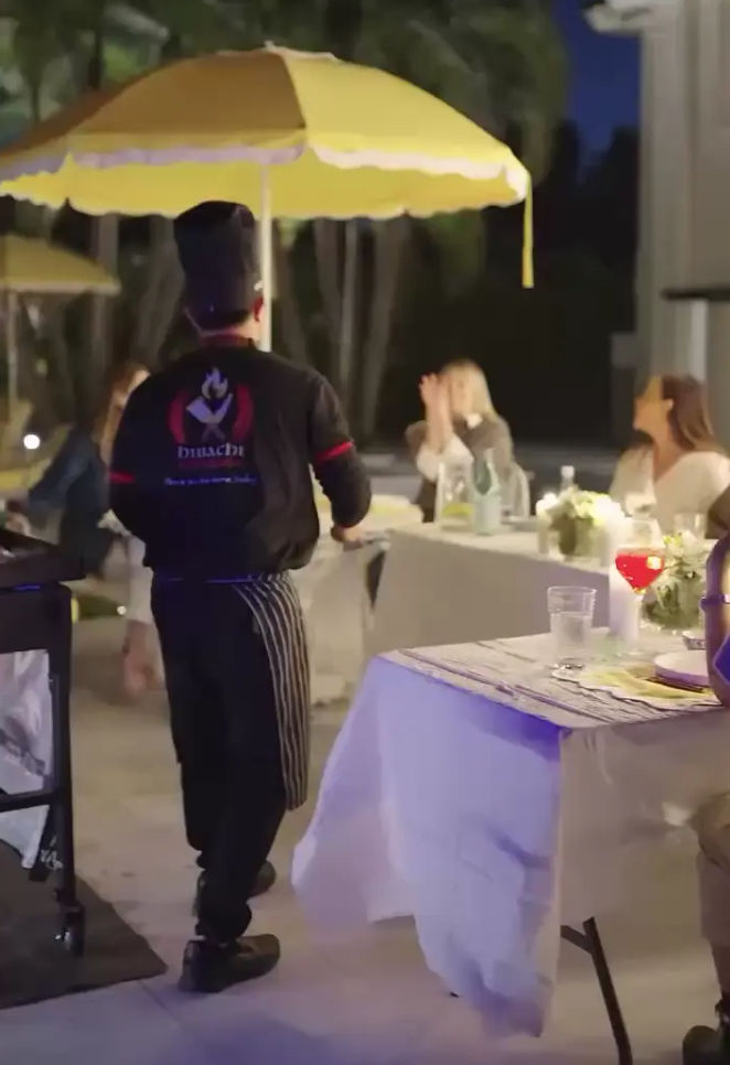 Server in a chef hat walking under a yellow patio umbrella at an al fresco evening dinner — outdoor patio dining with tables set, drinks and flower centerpieces.