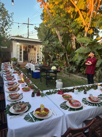 Sunset tropical backyard dinner party with long white-tablecloth tables, monstera leaf placemats, bowls and candles, string lights overhead, grill and chef in a burgundy jacket.