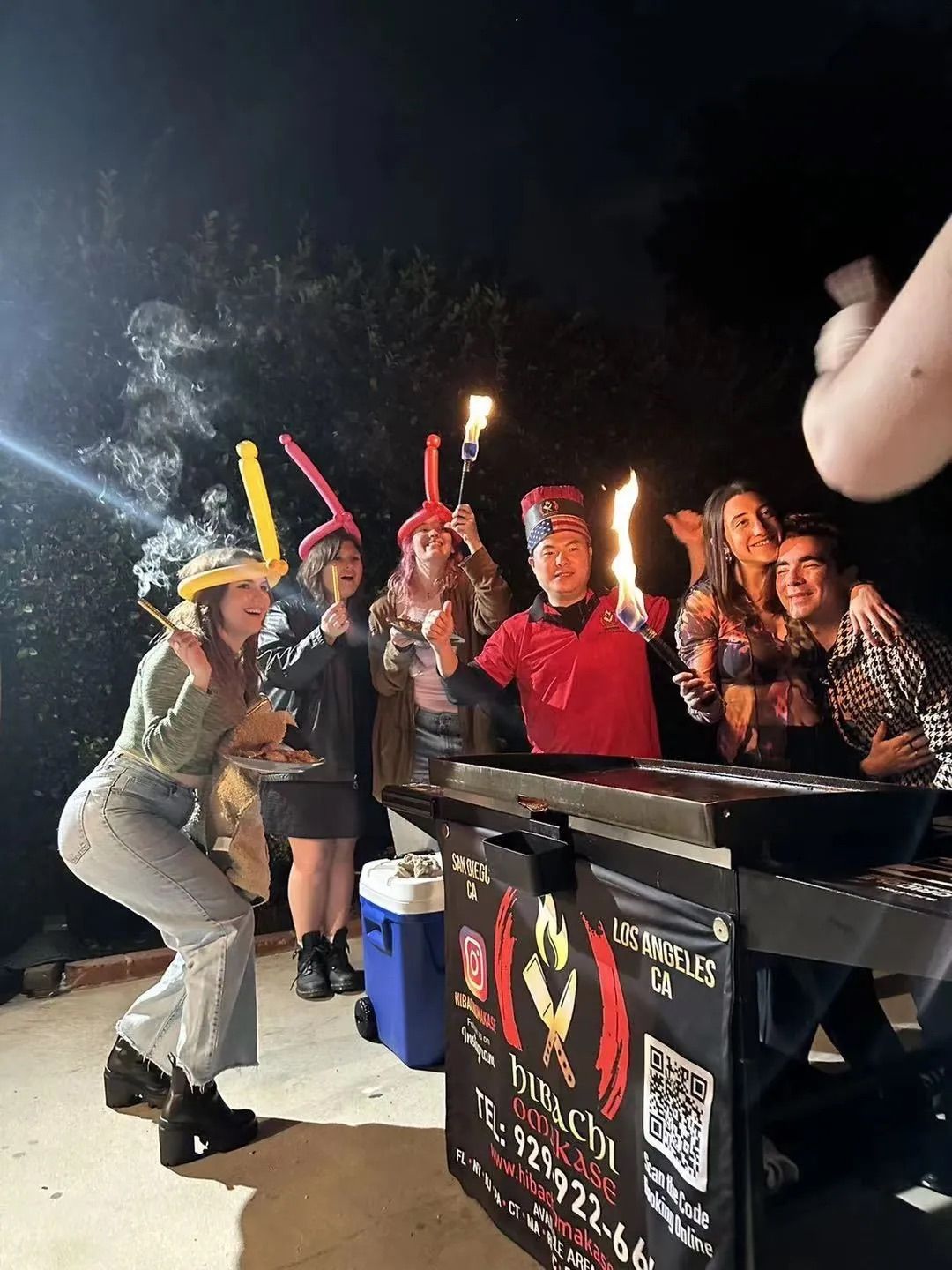 Nighttime Los Angeles backyard party: friends wearing balloon hats pose around an outdoor hibachi grill cart as a chef lights flaming torches.