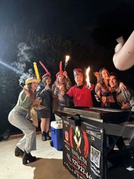 Outdoor nighttime hibachi party in Los Angeles — friends gathered around a portable grill with a chef holding flaming torches, guests wearing balloon hats and laughing.