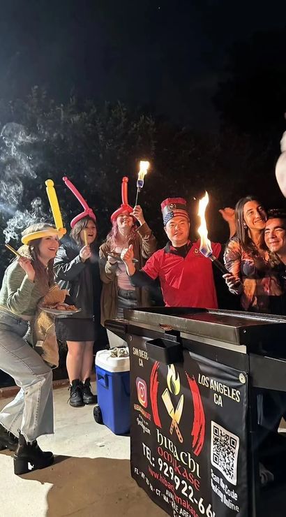 Nighttime outdoor hibachi celebration in Los Angeles — friends in balloon hats cheer with flaming skewers and a smiling chef by a grill.