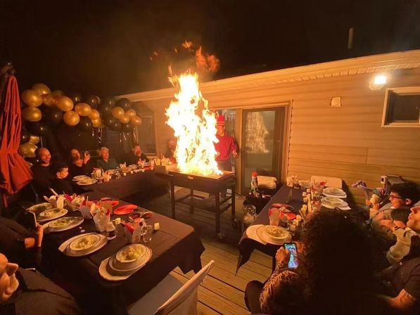 Nighttime backyard deck party with long tables and black-and-gold balloons; a cook creates a dramatic tall flame on a grill while seated guests watch, eat, and take photos.