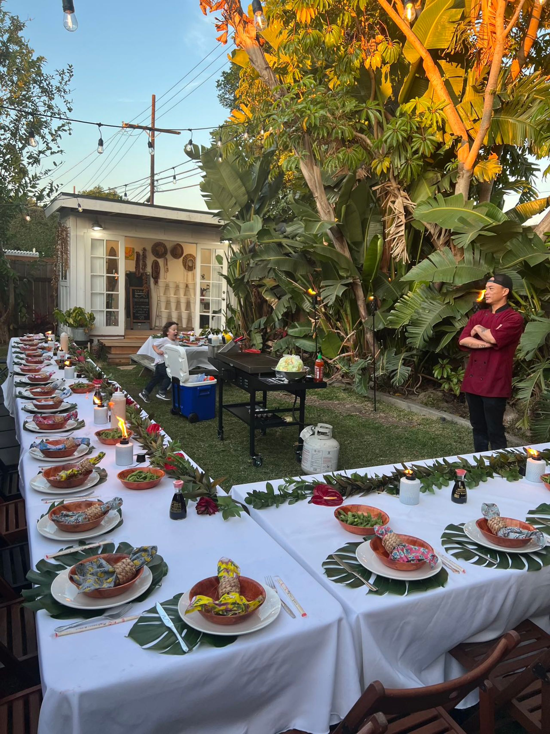 Backyard luau-style dinner in a lush tropical garden: long white-tablecloth tables with palm-leaf chargers, bowls holding wrapped mini pineapples, chopsticks, tiki torches and string lights; a chef stands by a grill near a small garden studio.