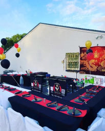 Outdoor hibachi-style patio party setup with U-shaped tables draped in black and red, round black plates and chopsticks at each place, central hibachi grills, dragon tapestry on the wall and black‑red‑gold balloons.