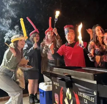 Group of people wearing balloon hats and party hats gathered around a tabletop grill at night, holding lit skewers and small torches in a festive outdoor Los Angeles celebration.