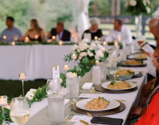 Outdoor tent wedding reception head table with white floral centerpieces, candlelight, greenery and plated rice entrees at elegant place settings