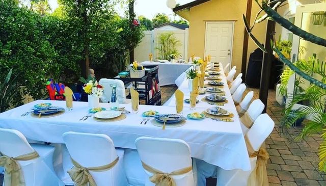 Outdoor backyard patio banquet with a long L-shaped table, white linens, gold-sashed chairs, blue plates and yellow folded napkins for a festive garden dining setup.