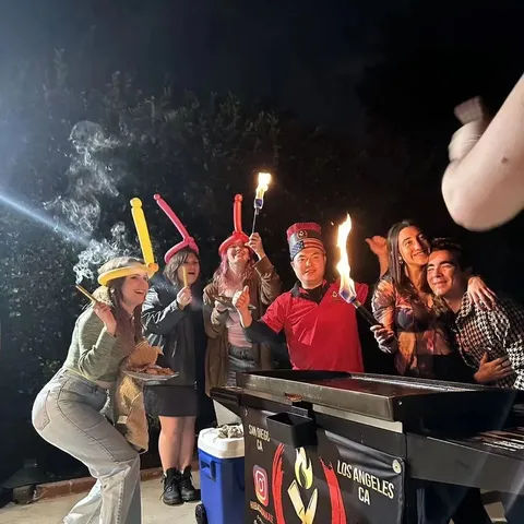 Friends at a nighttime Los Angeles backyard party around a mobile grill, wearing balloon hats and holding flaming torches and skewers while smiling for a photo.