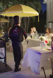 Chef in a striped apron carrying a bright yellow parasol walks between outdoor patio tables at an evening al fresco dinner party with guests at candlelit tables.