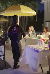 Evening outdoor patio dining scene with a server in a chef hat walking under a yellow umbrella toward guests seated at white tablecloths with glassware and a red cocktail.