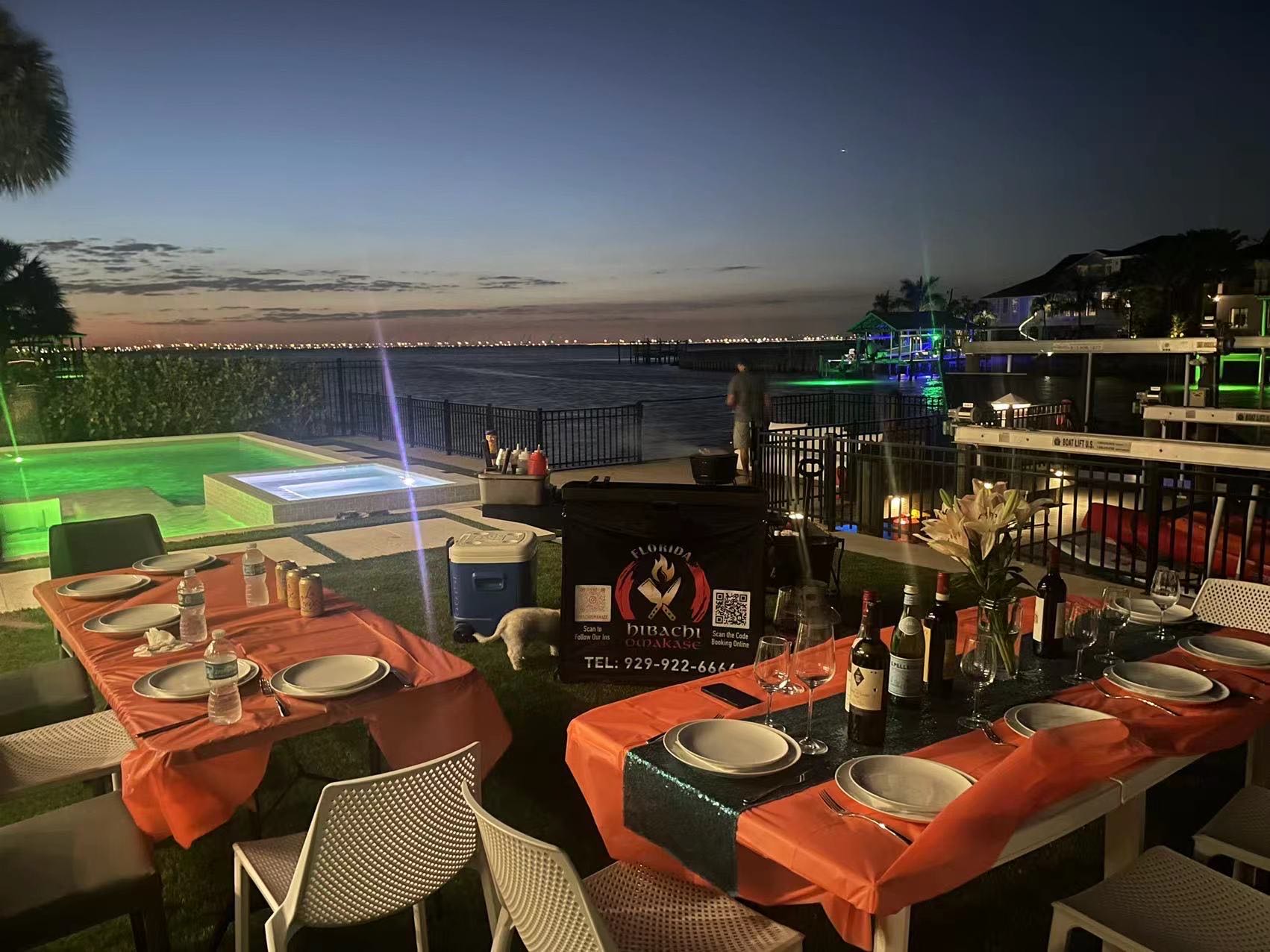 Cozy evening waterfront backyard dining at sunset with orange-covered tables, wine bottles, a lit pool and spa, and docks with bay lights in the background.