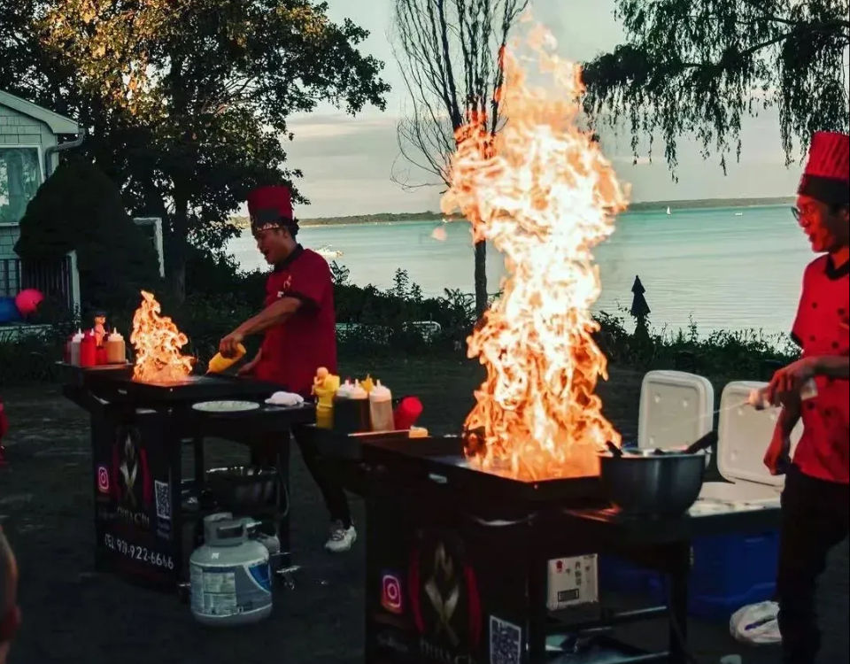 Two chefs in red uniforms performing a lakeside outdoor teppanyaki grill show at sunset, dramatic towering flames rising from flat-top griddles with condiments and coolers nearby.