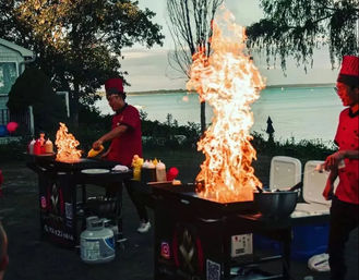 Two chefs in red uniforms performing a lakeside outdoor teppanyaki grill show at sunset, dramatic towering flames rising from flat-top griddles with condiments and coolers nearby.