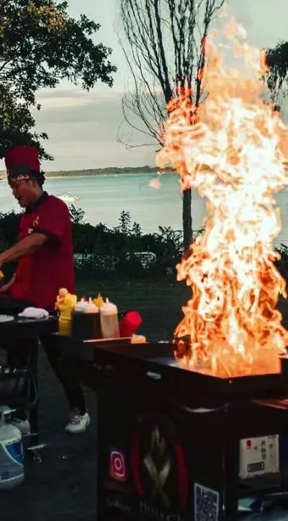 Two chefs in red uniforms performing a lakeside outdoor teppanyaki grill show at sunset, dramatic towering flames rising from flat-top griddles with condiments and coolers nearby.