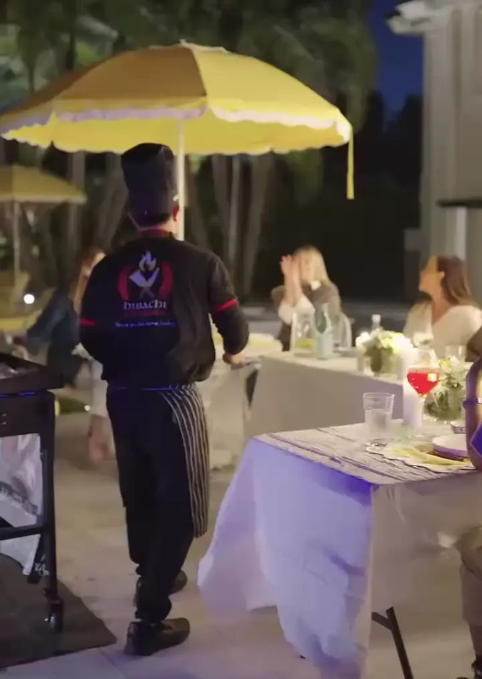 Outdoor evening dinner scene with a server in a striped apron walking beneath a yellow patio umbrella past white‑linened tables, floral centerpieces and seated guests.