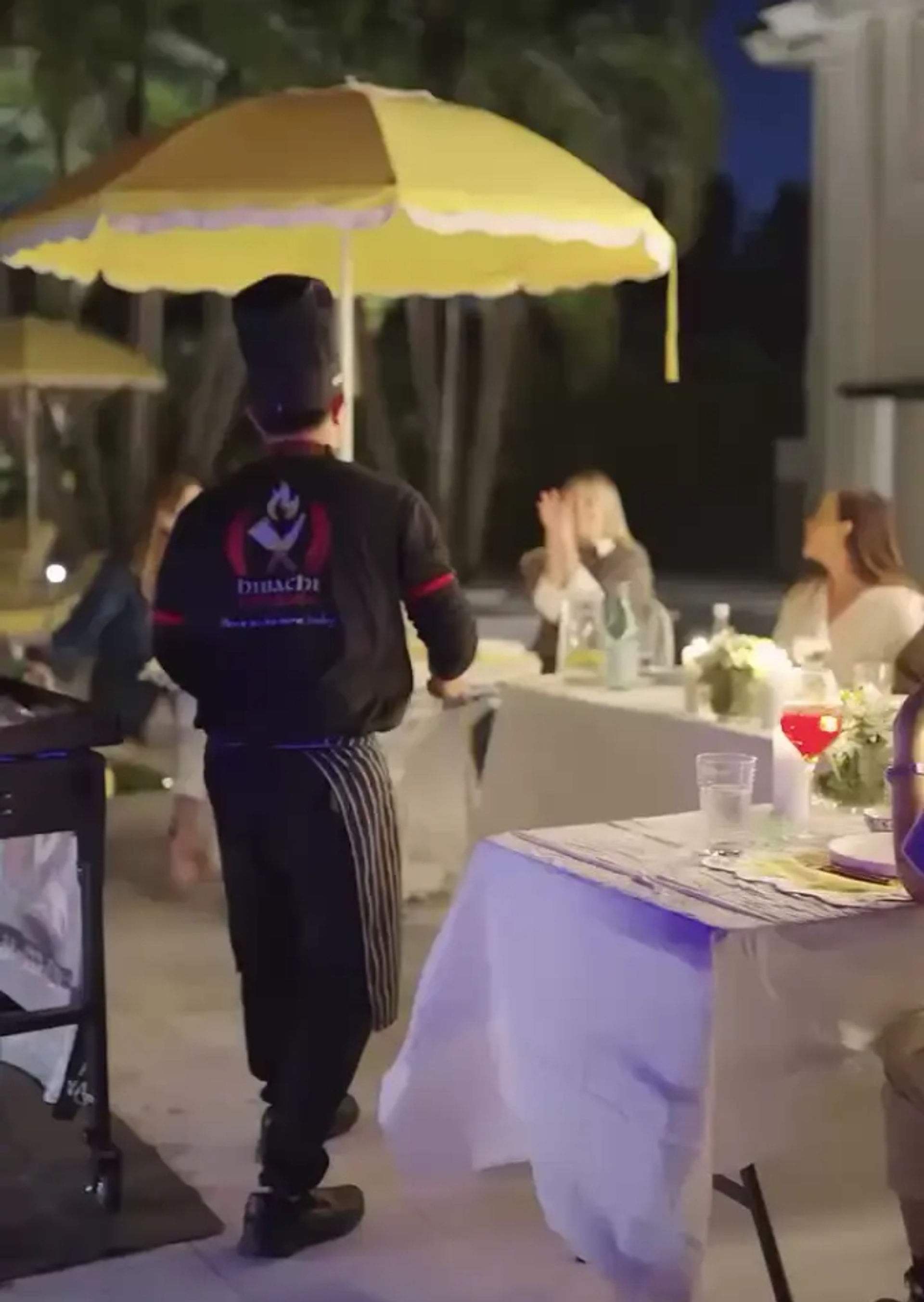Outdoor evening dinner scene with a server in a striped apron walking beneath a yellow patio umbrella past white‑linened tables, floral centerpieces and seated guests.
