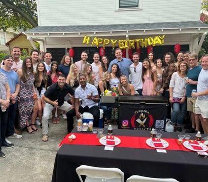 Backyard birthday party group photo: about 30 smiling adults in front of a suburban house under gold “HAPPY BIRTHDAY” balloons, with a hibachi-style grill station and red-and-black buffet tables set with plates and drinks.
