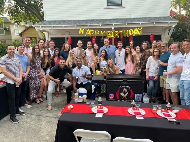 Backyard birthday party group photo: about 30 smiling adults in front of a suburban house under gold “HAPPY BIRTHDAY” balloons, with a hibachi-style grill station and red-and-black buffet tables set with plates and drinks.