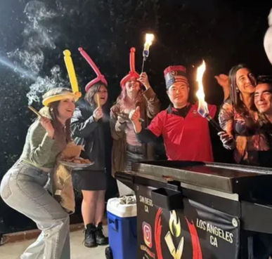 Smiling group celebrating at night in Los Angeles, wearing balloon hats and holding flaming torches next to a mobile food cart.