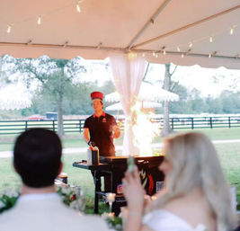 Live chef performing a flaming teppanyaki cooking show under a white tent at an outdoor country wedding reception, guests watching with string lights and a pasture fence in the background.