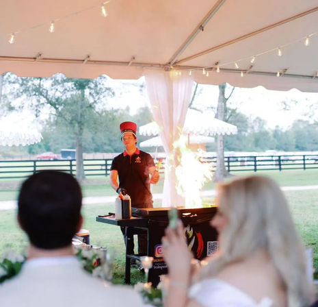 Live chef performing a flaming teppanyaki cooking show under a white tent at an outdoor country wedding reception, guests watching with string lights and a pasture fence in the background.