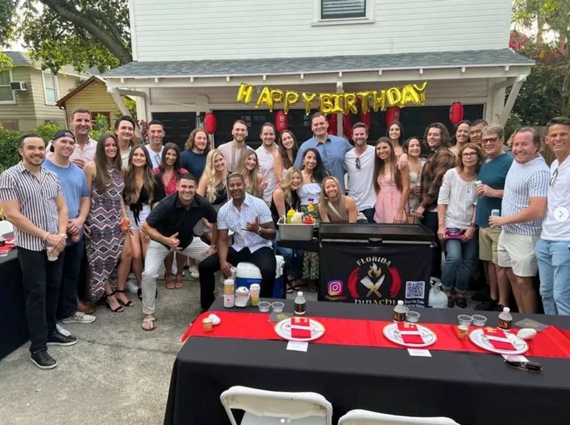 Large group of friends posing at a backyard birthday party in front of a house with gold "Happy Birthday" balloons, an outdoor grill station, red-and-black table settings and festive decorations.