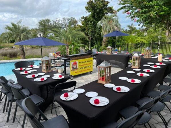 Poolside backyard party setup with U-shaped black-clothed tables and folding chairs, white plates topped with red napkin roses, wooden lantern centerpieces, blue umbrellas, palm trees and a swimming pool in the background.
