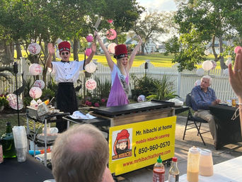 Backyard mobile hibachi teppanyaki: chef in white jacket and a birthday guest wearing a sash and tall red hat raise spatulas by a white picket fence and pink paper lanterns, guests seated at outdoor tables.