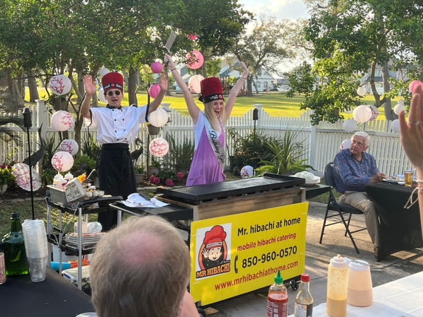 Backyard mobile hibachi teppanyaki: chef in white jacket and a birthday guest wearing a sash and tall red hat raise spatulas by a white picket fence and pink paper lanterns, guests seated at outdoor tables.