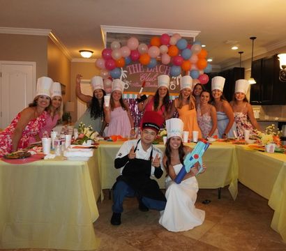 Group of women and a chef in a home kitchen celebrating under a colorful balloon arch, all wearing paper chef hats and smiling around long yellow-covered tables with plates and drinks, while a woman in a white dress kneels holding a blue water gun.