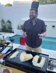 Smiling chef in a tall hat cooks heart- and U-shaped rice on a teppanyaki-style griddle at a poolside backyard patio