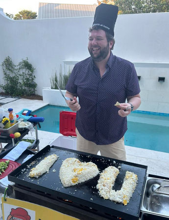 Smiling chef in a tall hat cooks heart- and U-shaped rice on a teppanyaki-style griddle at a poolside backyard patio