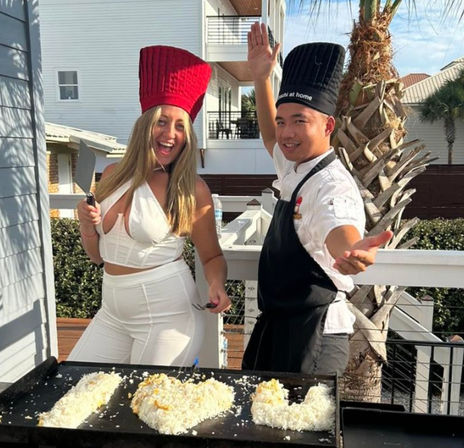 Smiling duo on a sunny beach-house balcony by a palm tree cooking at an outdoor teppanyaki grill — woman in white with a red chef hat and spatula and a chef in a black hat and apron shaping heart-shaped rice on the griddle.