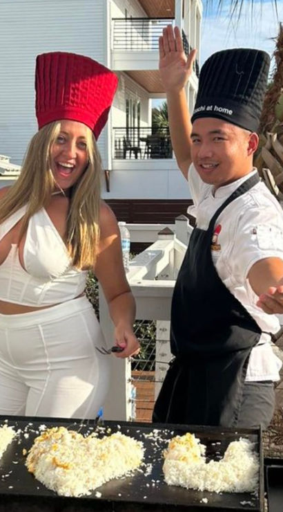 Smiling duo on a sunny beach-house balcony by a palm tree cooking at an outdoor teppanyaki grill — woman in white with a red chef hat and spatula and a chef in a black hat and apron shaping heart-shaped rice on the griddle.