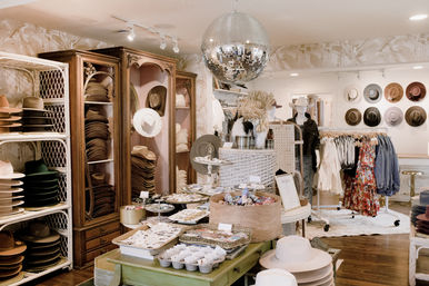Boho-chic women's boutique interior with shelves of hats, racks of dresses and denim, accessory-strewn tables, dried floral displays and a hanging disco ball.