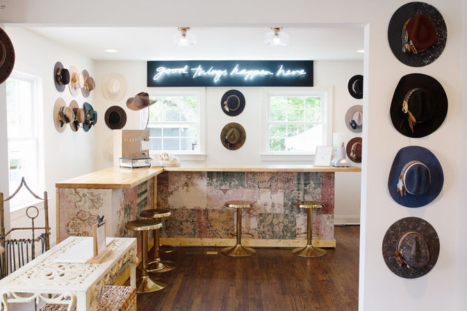 Bright, airy hat boutique interior with hats mounted on white walls, a wood-topped counter with patterned front, three brass bar stools, and a glowing neon sign reading "good things happen here".