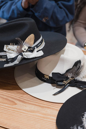 Two stylish wide-brim felt fedoras, one black and one cream, trimmed with ribbons, feathers and decorative pins, displayed on a wooden table.