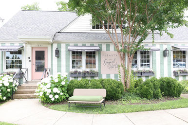 Charming pastel-striped boutique storefront with a pink door, black awnings, window flower boxes, white hydrangeas, a bench and front tree