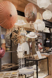 Boutique hat display with stylish wide-brim hats on a sculptural stand, hanging paper parasols, crystal globe chandeliers, and mirrored vintage accents in a retail interior.