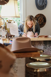 Woman leaning over a blush felt fedora on a wooden counter in a cozy hat boutique, surrounded by displayed hats, brass stools and a warm table lamp.