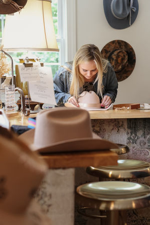 Woman leaning over a blush felt fedora on a wooden counter in a cozy hat boutique, surrounded by displayed hats, brass stools and a warm table lamp.